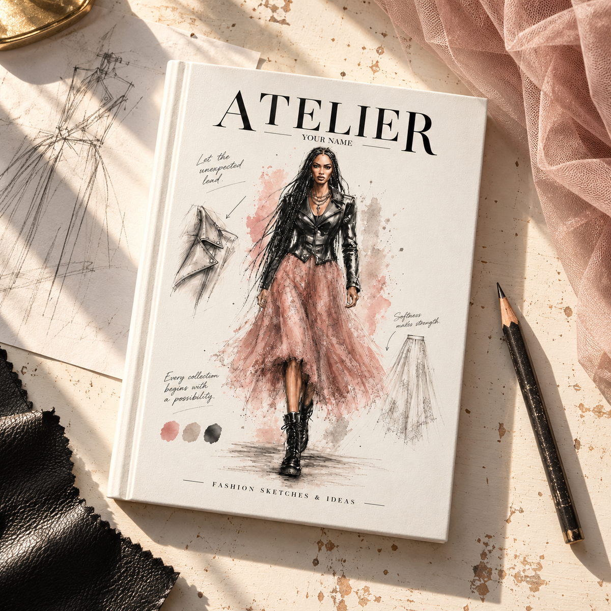 A hardcover sketchbook titled 'ATELIER' lies open on a styled flat-lay surface with fashion sketches, a pencil, black leather fabric, and pink tulle. The cover features a fashion illustration of a woman in a black top and pink skirt.