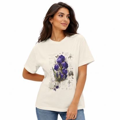 A woman wearing a cream-colored short-sleeve t-shirt with a purple iris and botanical sketch graphic on the front, paired with blue jeans, set against a plain white background.